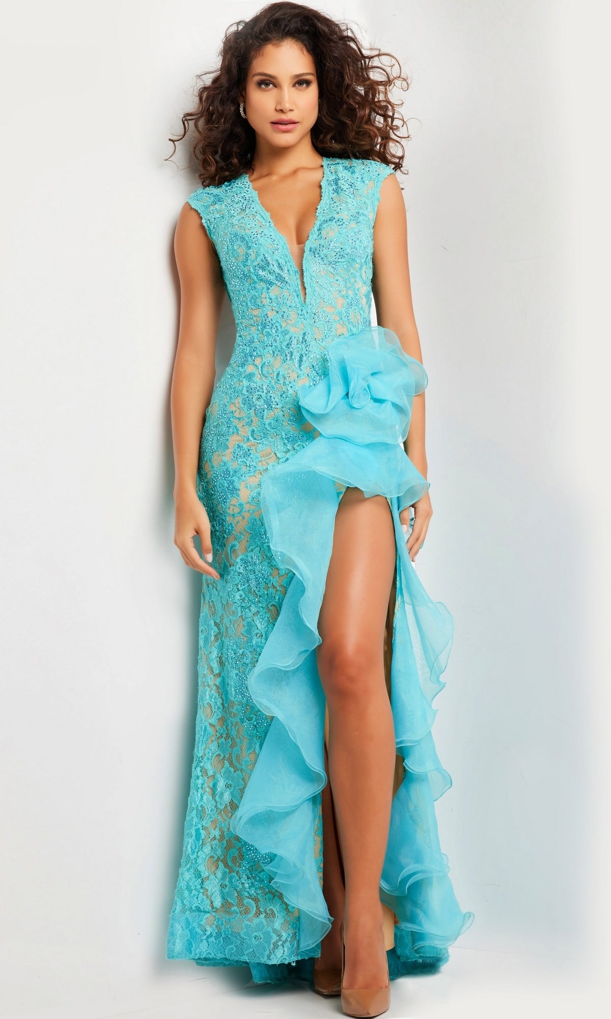 A woman wearing the Jovani Ruffled Long Lace Prom Dress 38668 in turquoise, with a plunging neckline, large floral accent, and thigh-high slit, stands against a plain light background.